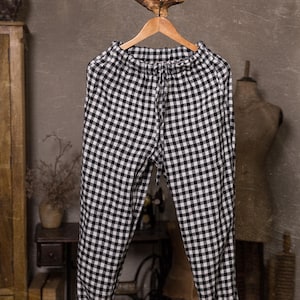 Linen pants in black and white check, displayed on a wooden hanger in a full-length front view, with a gathered elastic waistband, drawstring ties, loose tapered legs, turned-up hems, and a rustic indoor background.