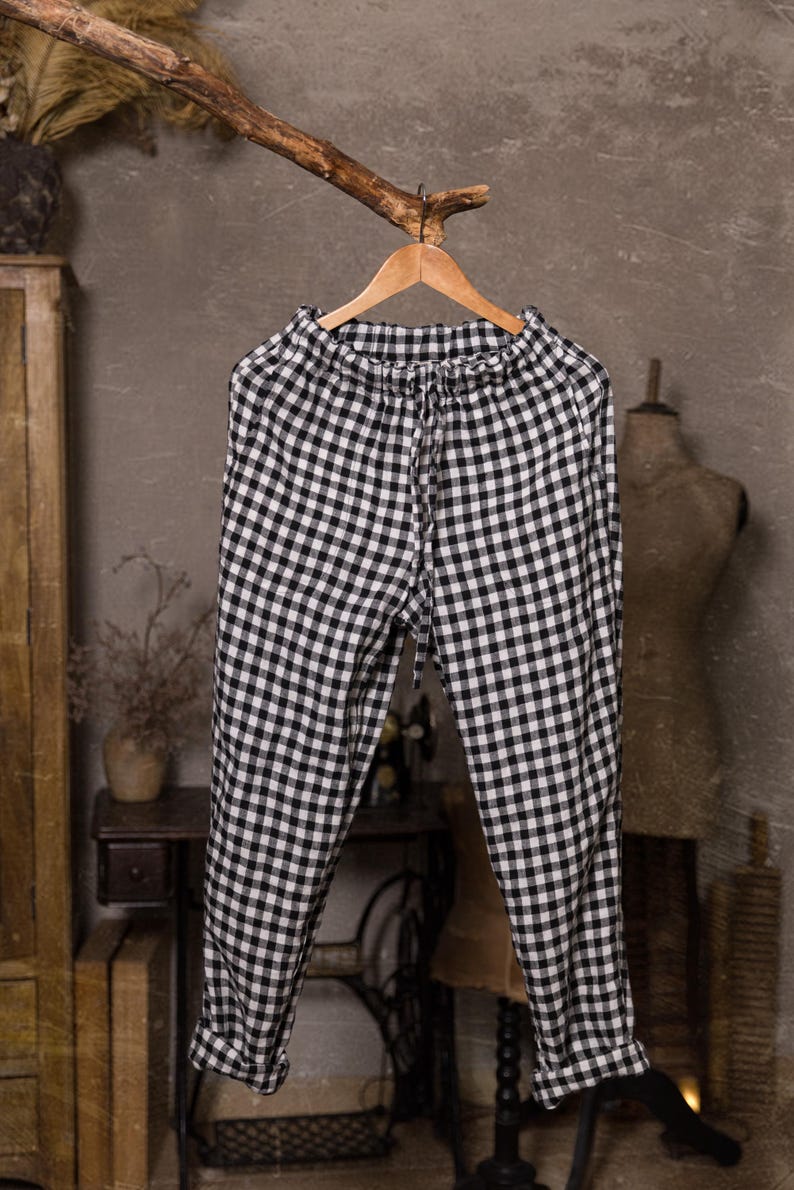 Linen pants in black and white check, displayed on a wooden hanger in a full-length front view, with a gathered elastic waistband, drawstring ties, loose tapered legs, turned-up hems, and a rustic indoor background.
