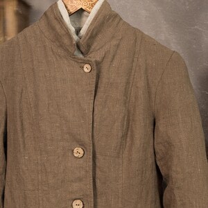 Brown linen jacket close-up on a wooden hanger, front view, with natural-tone buttons, high folded collar, vertical panel seams, textured linen fabric, and pale linen neckline showing inside.