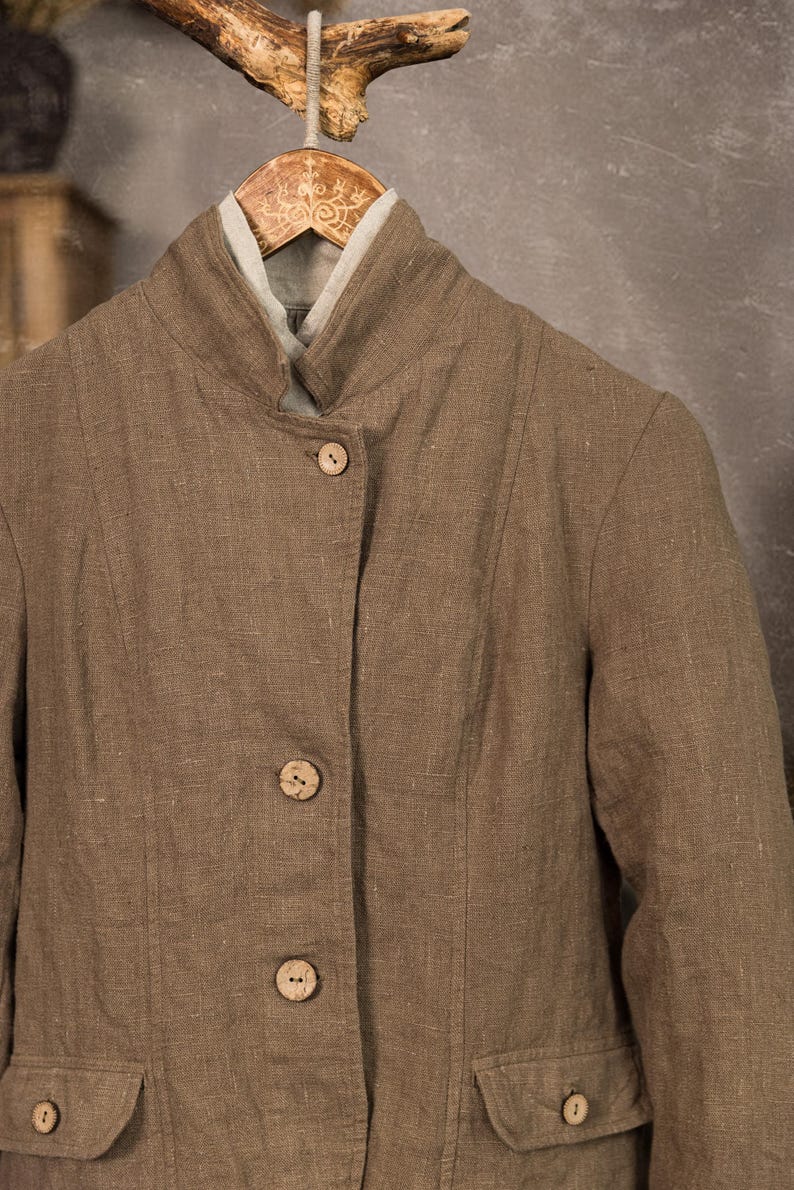 Brown linen jacket close-up on a wooden hanger, front view, with natural-tone buttons, high folded collar, vertical panel seams, textured linen fabric, and pale linen neckline showing inside.