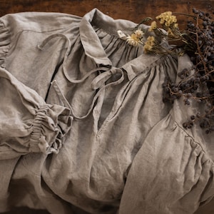 Close-up flat lay of one natural flax linen dress showing tie-front neckline, gathered collar, and long sleeve with ruffled cuff, on a wooden surface with dried berries and flowers.
