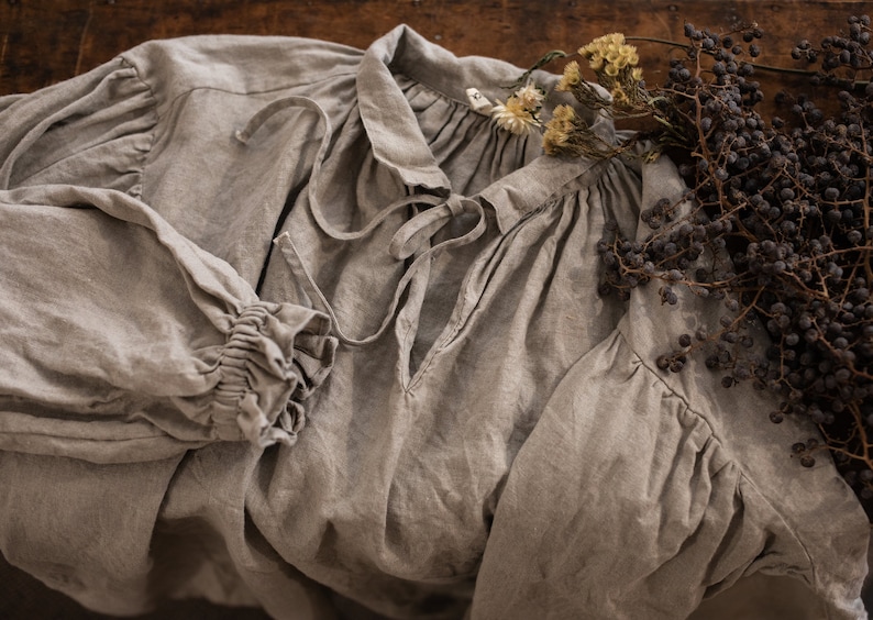 Close-up flat lay of one natural flax linen dress showing tie-front neckline, gathered collar, and long sleeve with ruffled cuff, on a wooden surface with dried berries and flowers.