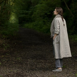 Natural linen dress coat with long sleeves, button front, and two side pockets, worn by a person standing on a stone-covered forest path. Styled with a large linen scarf, gray pants, and gray shoes; background includes dense greenery.