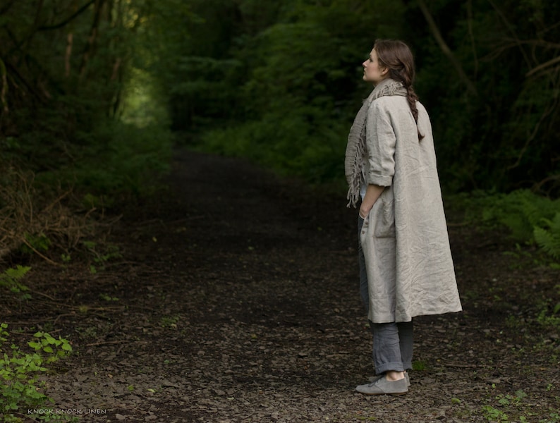 Natural linen dress coat with long sleeves, button front, and two side pockets, worn by a person standing on a stone-covered forest path. Styled with a large linen scarf, gray pants, and gray shoes; background includes dense greenery.