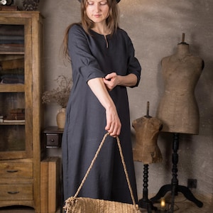 Front view of a model in a maxi linen dress in deep navy with 3/4 sleeves and relaxed fit, sleeves pushed up, holding a woven straw bag in front of her in a neutral rustic room.