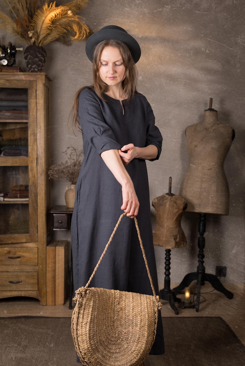 Front view of a model in a maxi linen dress in deep navy with 3/4 sleeves and relaxed fit, sleeves pushed up, holding a woven straw bag in front of her in a neutral rustic room.