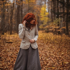 A light beige linen blazer with a button closure and a long grey skirt. The outfit is worn in a forest setting with autumn leaves on the ground. The person has red hair.