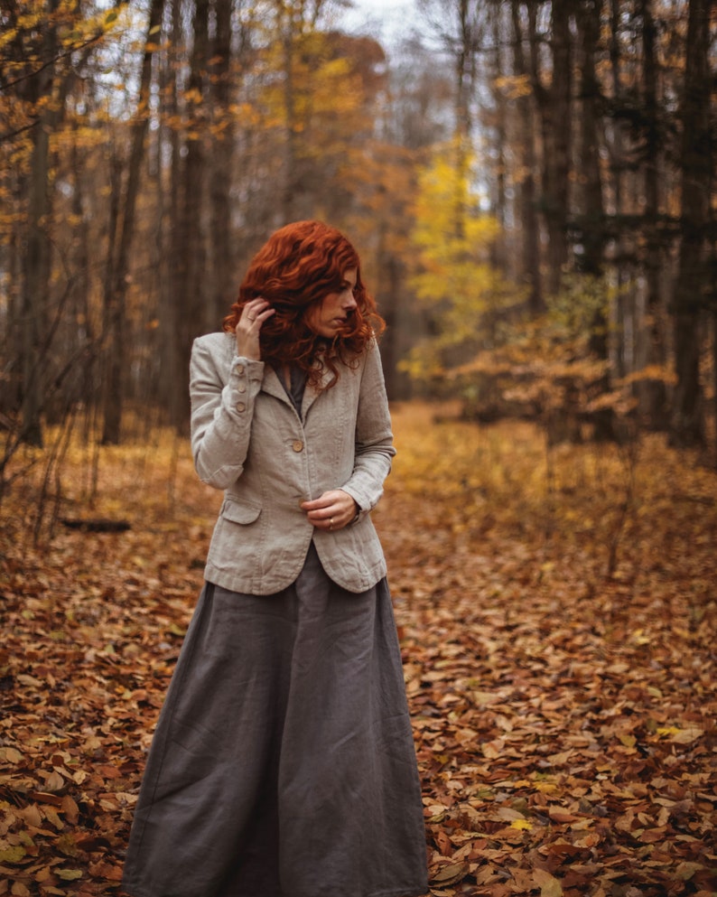 A light beige linen blazer with a button closure and a long grey skirt. The outfit is worn in a forest setting with autumn leaves on the ground. The person has red hair.