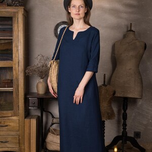 One maxi summer linen dress in deep navy shown on a model indoors, ankle length with 3/4 sleeves and simple kaftan silhouette, worn with a dark hat and brown shoes in a rustic studio setting.