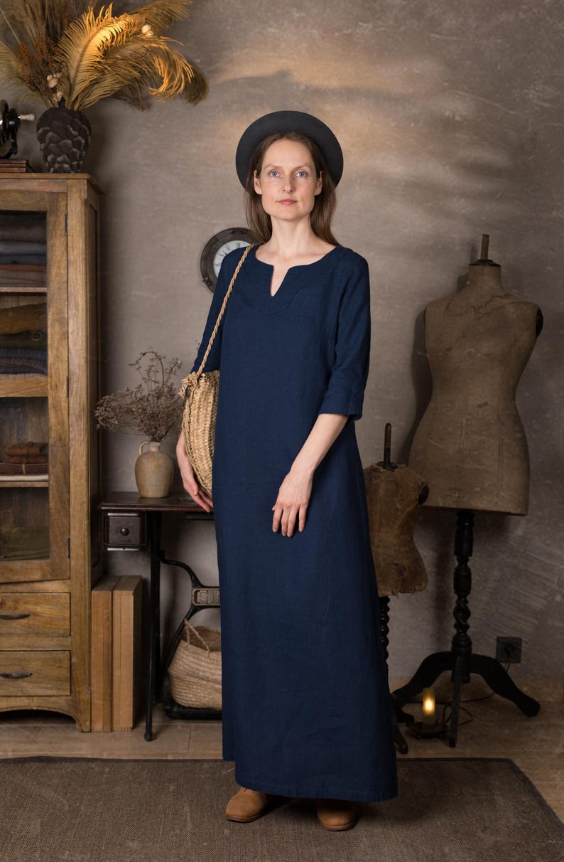 One maxi summer linen dress in deep navy shown on a model indoors, ankle length with 3/4 sleeves and simple kaftan silhouette, worn with a dark hat and brown shoes in a rustic studio setting.