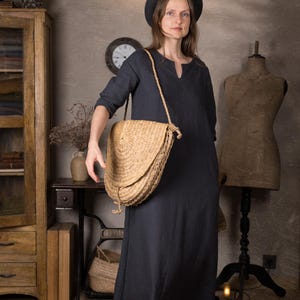 Front view of a woman in a long dark blue linen dress with 3/4 sleeves and relaxed fit, styled with a round black hat and straw crossbody bag, standing in a rustic indoor studio setting.