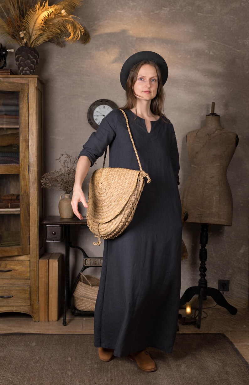 Front view of a woman in a long dark blue linen dress with 3/4 sleeves and relaxed fit, styled with a round black hat and straw crossbody bag, standing in a rustic indoor studio setting.
