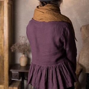 One purple linen blouse with long sleeves and peplum ruffle at the hips, shown from the back on a model standing in a rustic studio.