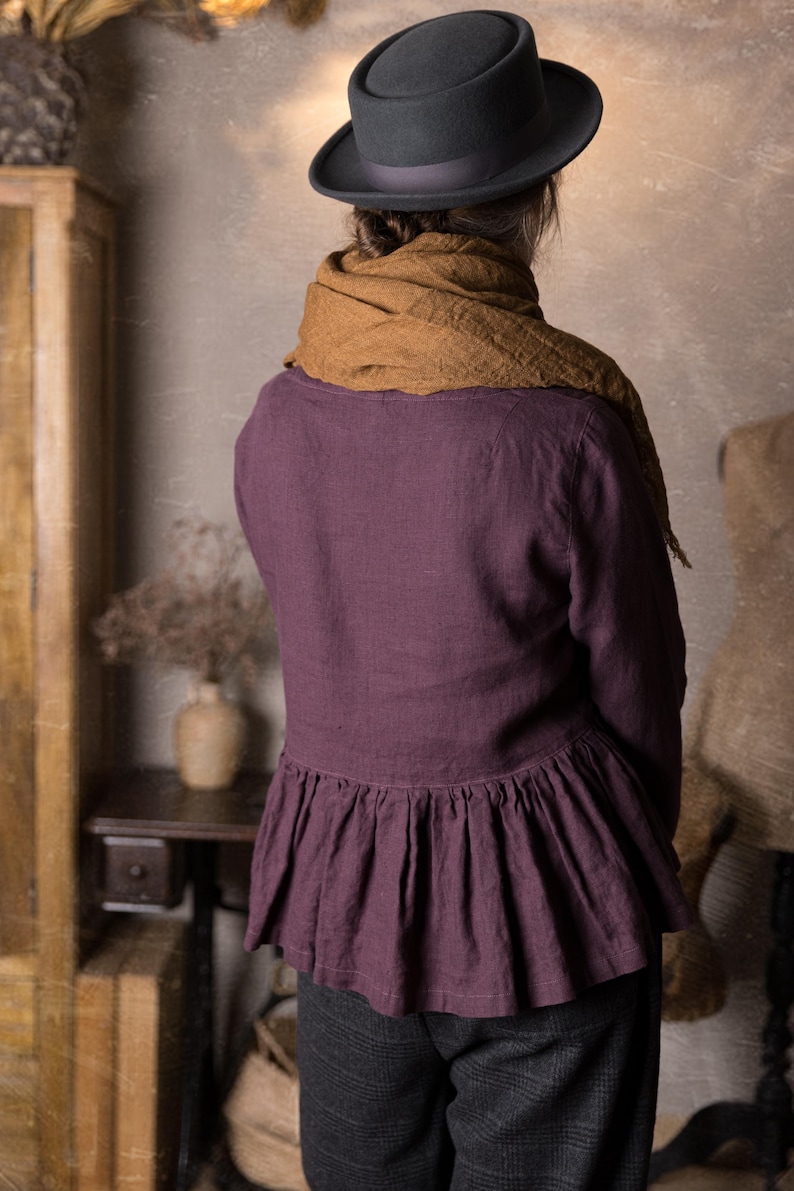 One purple linen blouse with long sleeves and peplum ruffle at the hips, shown from the back on a model standing in a rustic studio.