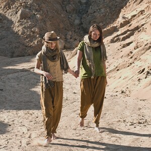 Two people walking barefoot on rocky terrain, holding hands. Both wear loose, earth-toned linen outfits with scarves around their necks. Scene is sunlit with deep shadows, suggesting late afternoon. Linen top, loose linen shirt, buttoned shirt.