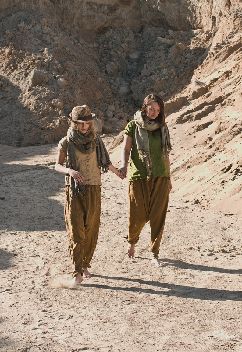 Two people walking barefoot on rocky terrain, holding hands. Both wear loose, earth-toned linen outfits with scarves around their necks. Scene is sunlit with deep shadows, suggesting late afternoon. Linen top, loose linen shirt, buttoned shirt.