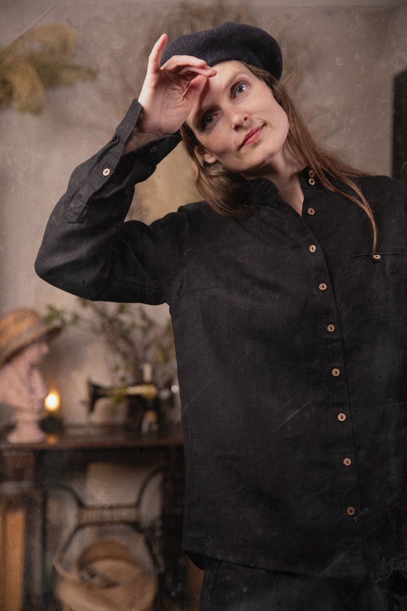 Person wearing a black linen button-down shirt Village, waist-up front view, stand collar, long sleeves, row of light buttons, left chest pocket with button, hand raised to touch a black beret, blurred sewing machine table behind.
