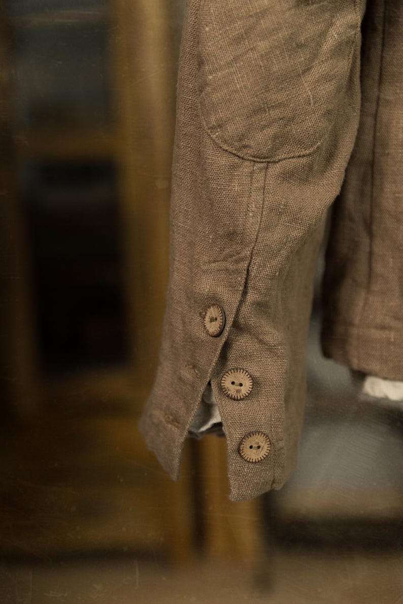 Detail of a brown linen jacket sleeve. The sleeve has three wooden buttons and a rounded elbow patch. The fabric has a textured look, and the jacket appears to be tailored.