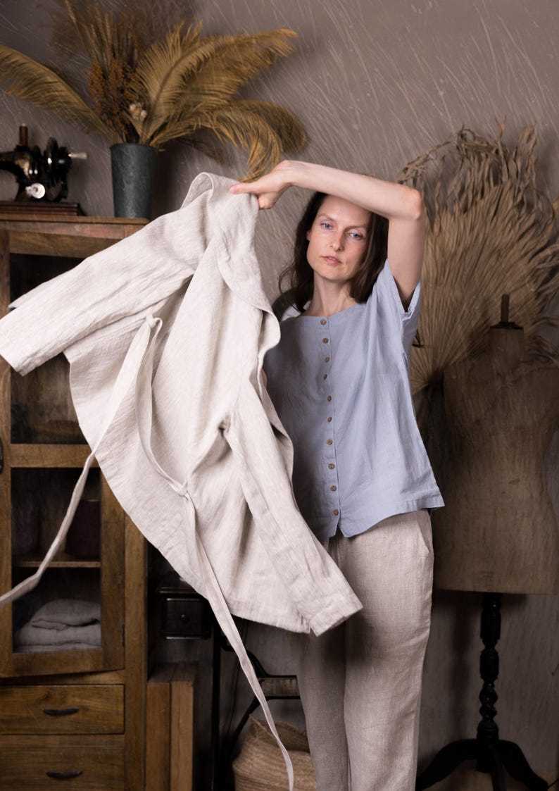 Person indoors putting on a natural linen jacket over a dusty sky blue short-sleeved linen top. Only upper body visible. Background includes wooden cabinet with folded linens and sewing tools. Buttoned linen top, linen blouse, loose linen shirt.