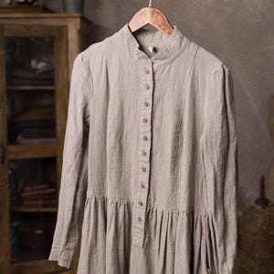 natural flax linen button-front dress displayed on hanger from a tree branch, front view cropped at mid-skirt, stand collar, row of buttons to the waist, long sleeves, gathered lower panel, green scissors at right side.