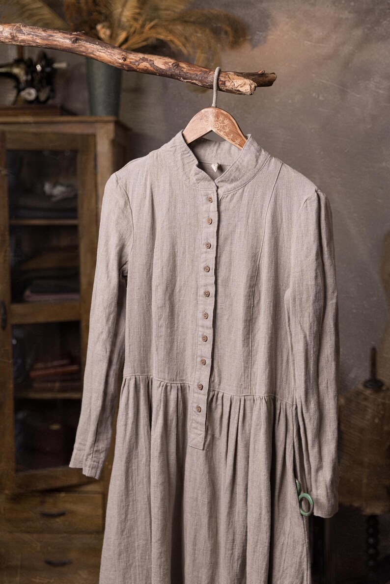 natural flax linen button-front dress displayed on hanger from a tree branch, front view cropped at mid-skirt, stand collar, row of buttons to the waist, long sleeves, gathered lower panel, green scissors at right side.