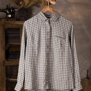 Gray and white checkered linen button-up shirt displayed on hanger, front full view, collar, long sleeves, center button placket with brown buttons, one chest patch pocket, slightly rounded hem; grey wall studio background with cabinet, knockknocklinen.