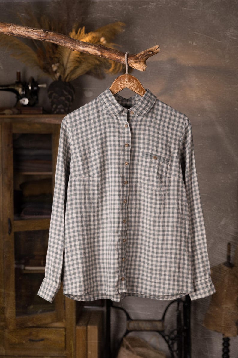 Gray and white checkered linen button-up shirt displayed on hanger, front full view, collar, long sleeves, center button placket with brown buttons, one chest patch pocket, slightly rounded hem; grey wall studio background with cabinet, knockknocklinen.