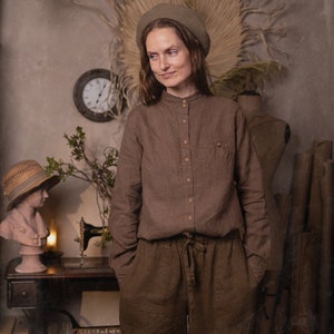 Waist-up photo of a model wearing a brown linen button-down shirt with stand collar, long sleeves, light buttons, and chest pocket, beige beret visible, mannequin and rolled paper in background, knockknocklinen.