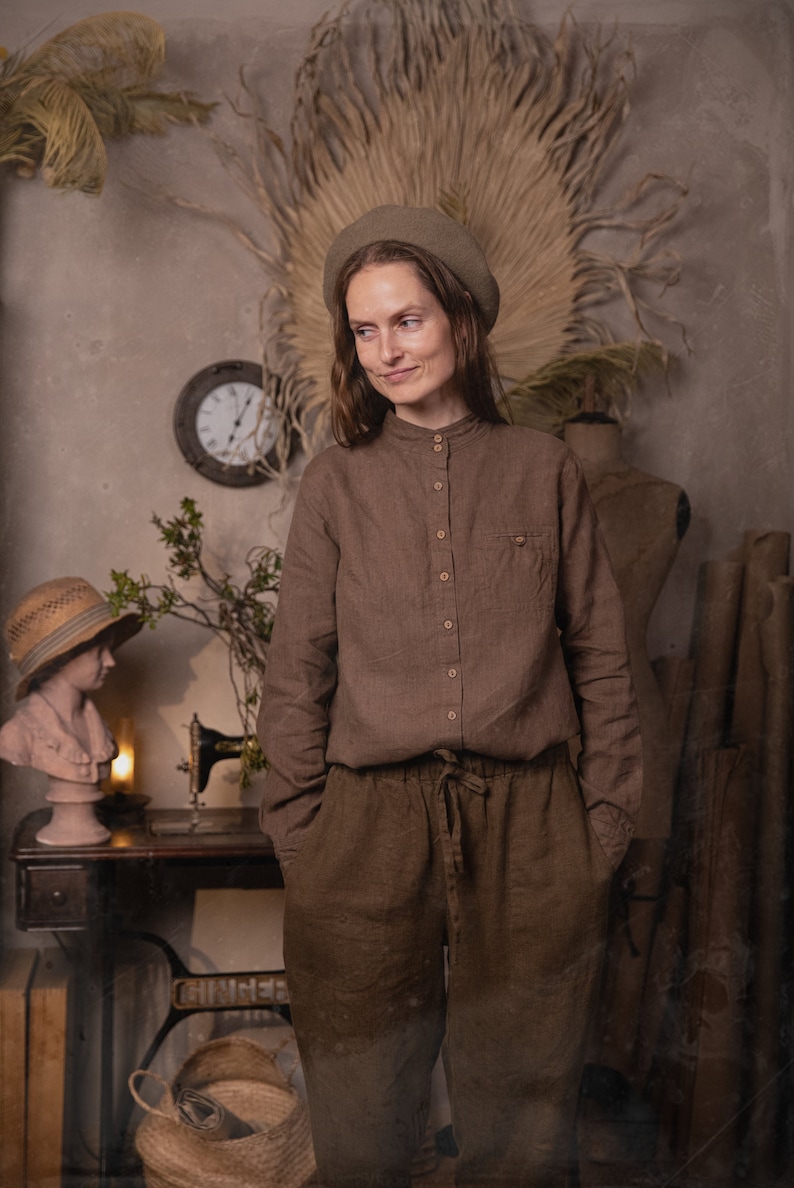 Waist-up photo of a model wearing a brown linen button-down shirt with stand collar, long sleeves, light buttons, and chest pocket, beige beret visible, mannequin and rolled paper in background, knockknocklinen.