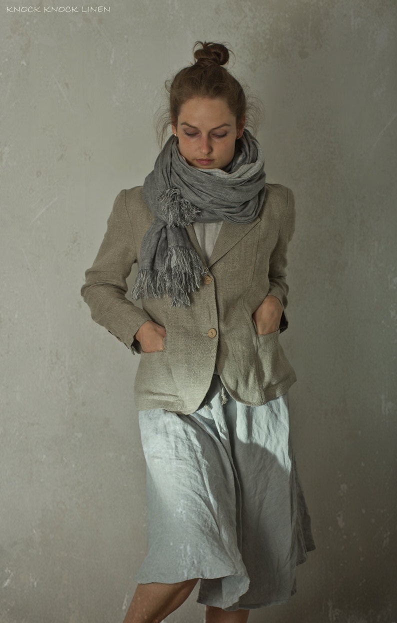 Grey-brown linen jacket worn by a model in a front full-length crop, with notched lapels, two visible buttons, large front pockets, long sleeves, and a grey scarf wrapped at the neck.