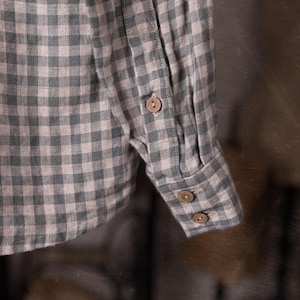 Close-up of a gingham linen shirt sleeve and cuff, showing buttoned cuff tab, two cuff buttons, placket seam, and creased fabric, shot as a detail while hanging, blurred studio props behind, knockknocklinen.