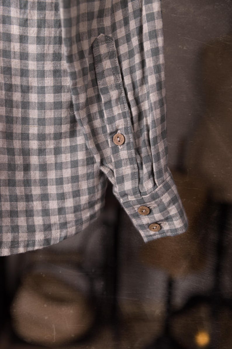 Close-up of a gingham linen shirt sleeve and cuff, showing buttoned cuff tab, two cuff buttons, placket seam, and creased fabric, shot as a detail while hanging, blurred studio props behind, knockknocklinen.