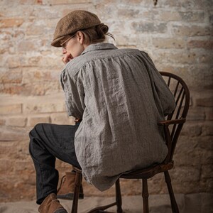 Side view on model wearing black linen pants, relaxed linen pants with tapered lower legs and natural creasing, photographed indoors with a wooden chair and brick wall.