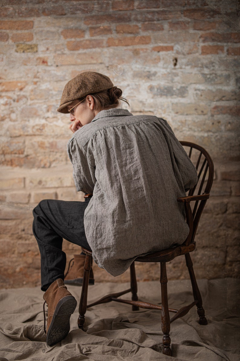 Side view on model wearing black linen pants, relaxed linen pants with tapered lower legs and natural creasing, photographed indoors with a wooden chair and brick wall.