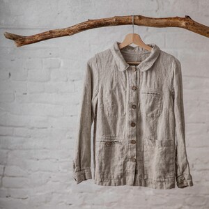 A natural flax linen shirt with a Peter pan collar, button-down front, and two front pockets, displayed hanging on a wooden hanger from a branch against a white brick wall. The loose-fitting workwear jacket features long sleeves and a natural fabric texture.