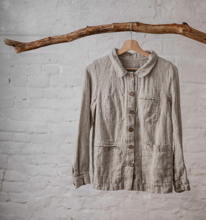 A natural flax linen shirt with a Peter pan collar, button-down front, and two front pockets, displayed hanging on a wooden hanger from a branch against a white brick wall. The loose-fitting workwear jacket features long sleeves and a natural fabric texture.