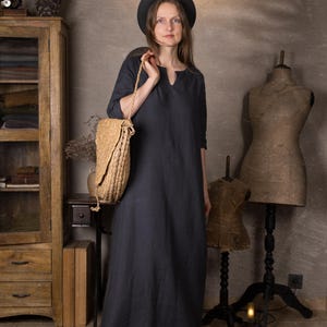 One model wearing a dark navy maxi linen dress with 3/4 sleeves and a split neckline, standing indoors in a rustic studio, holding a large woven straw shoulder bag and wearing a black hat.