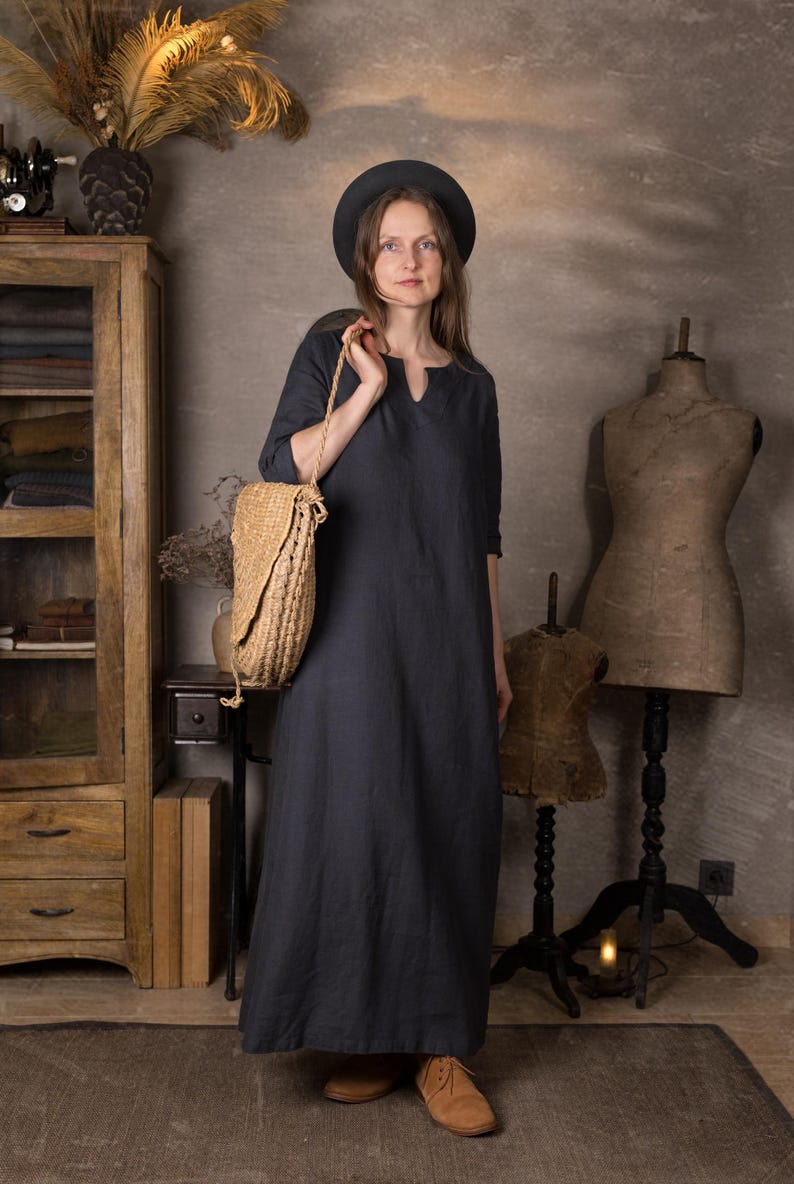 One model wearing a dark navy maxi linen dress with 3/4 sleeves and a split neckline, standing indoors in a rustic studio, holding a large woven straw shoulder bag and wearing a black hat.