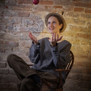 Linen pants in dark brown, shown on a seated model in a full-length three-quarter view, with a gathered waistband, visible drawstring ties, loose tapered legs, softly wrinkled fabric, and brown lace-up shoes, Knock Knock Linen.