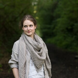 Natural linen buttoned dress coat with long sleeves, side pockets, and a loose silhouette, displayed worn by a person standing on a forest path. Styled with a large linen scarf; background includes greenery and soft natural light.