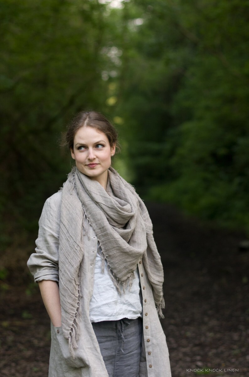 Natural linen buttoned dress coat with long sleeves, side pockets, and a loose silhouette, displayed worn by a person standing on a forest path. Styled with a large linen scarf; background includes greenery and soft natural light.