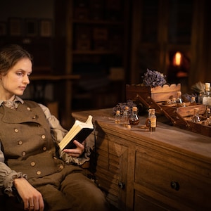 SPARROW linen vest in brown, shown on a seated model in a front three-quarter mid-length photo, with a sleeveless cut, two rows of light buttons, a curved overlapping front, over a light gray blouse, holding an open black book, Knock Knock Linen.