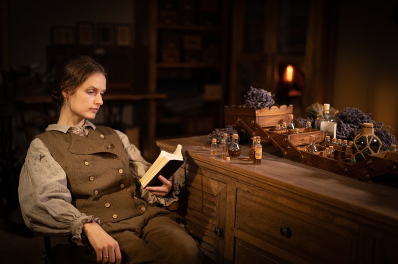 SPARROW linen vest in brown, shown on a seated model in a front three-quarter mid-length photo, with a sleeveless cut, two rows of light buttons, a curved overlapping front, over a light gray blouse, holding an open black book, Knock Knock Linen.