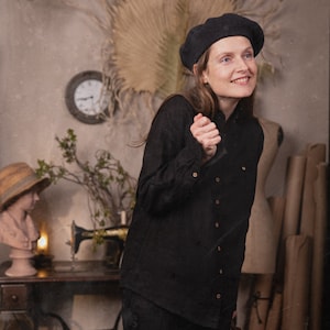 Three-quarter full-body photo of a person in a black linen shirt VILLAGE with long sleeves and light buttons, wearing a black beret and leaning forward, background shows a sewing machine table, bust with straw hat, and rolled paper tubes, knockknocklinen.