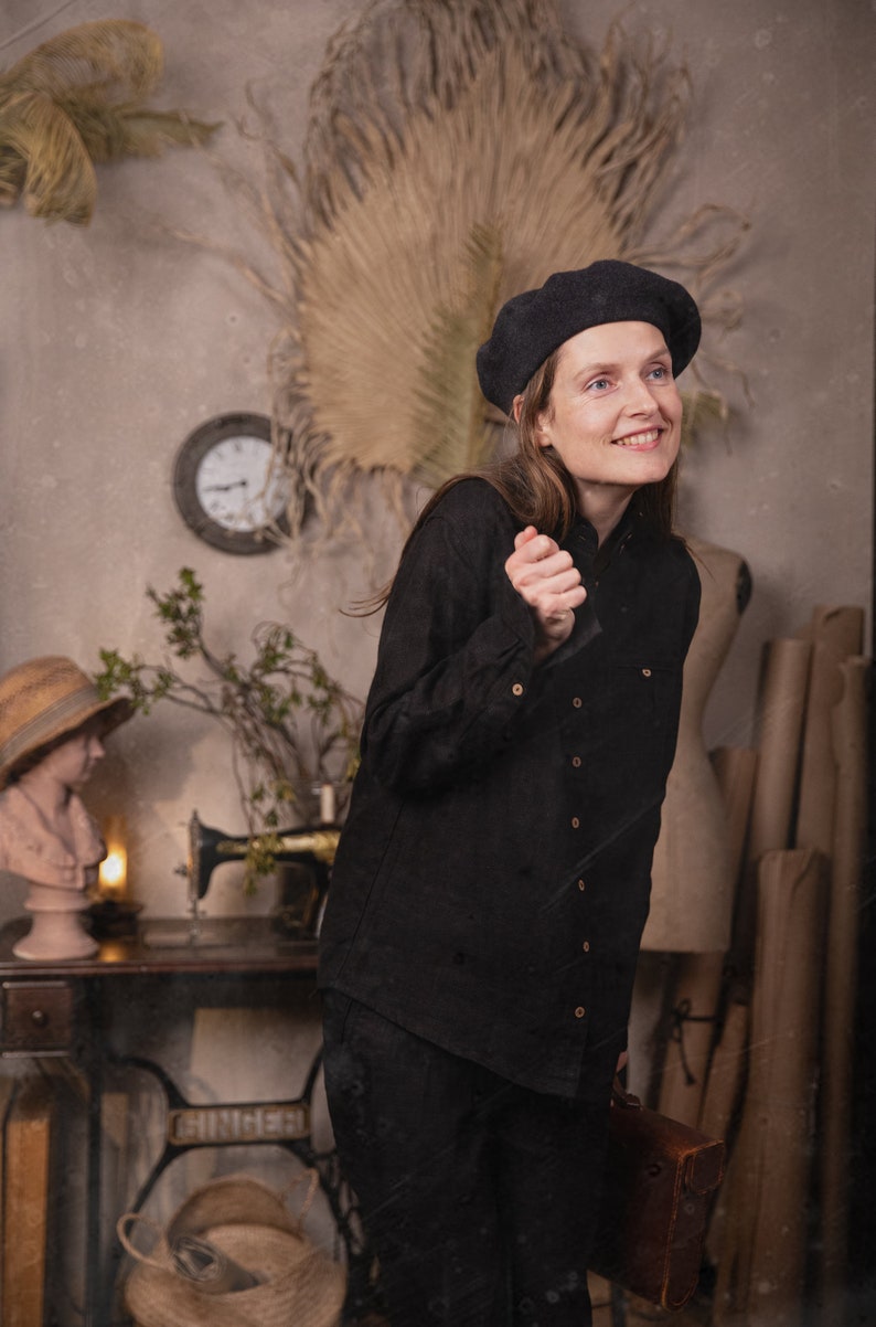 Three-quarter full-body photo of a person in a black linen shirt VILLAGE with long sleeves and light buttons, wearing a black beret and leaning forward, background shows a sewing machine table, bust with straw hat, and rolled paper tubes, knockknocklinen.