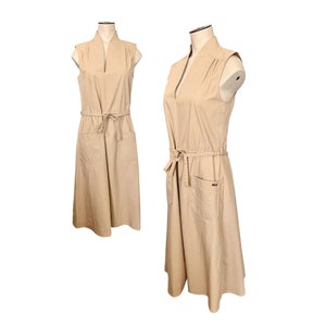 May include: A sleeveless, beige, cotton dress with a V-neckline, a tie belt, and two pockets. The dress is on a white background.