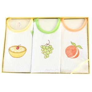May include: Set of three white terry cloth bibs with colorful fruit designs. The bibs have yellow, green, and orange trim. The designs include a lemon, grapes, and a peach.
