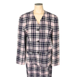 May include: A two-piece plaid suit in pink, navy, and white. The blazer has a V-neck, three buttons, and two front pockets. The skirt is visible at the bottom. The suit is displayed on a mannequin.