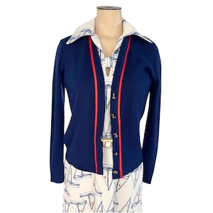 May include: A navy blue cardigan with red trim and gold buttons, layered over a white and blue patterned top. The top features a collared neckline and a sailboat print. The cardigan has long sleeves and a V-neck.