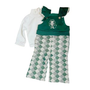 May include: A white turtleneck long-sleeve shirt and a green and white checkered overall set. The overall top is green with a lion design and ruffle detailing. The pants are a green and white checkered pattern.